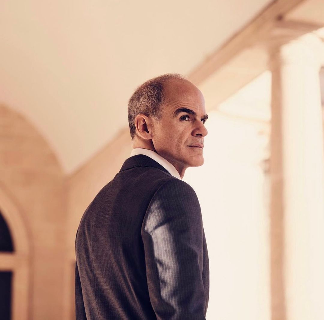 Adult male in a suit gazes over his shoulder indoors, standing near an arched walkway with columns; soft, neutral colors and diffused light.