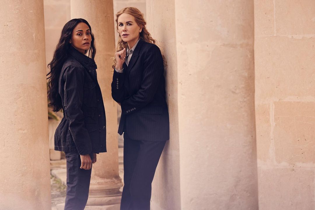 Two women, Zoe Saldana and Nicole Kidman, stand near stone columns, wearing dark jackets and pants, neutral expressions, in a medium outdoor shot.