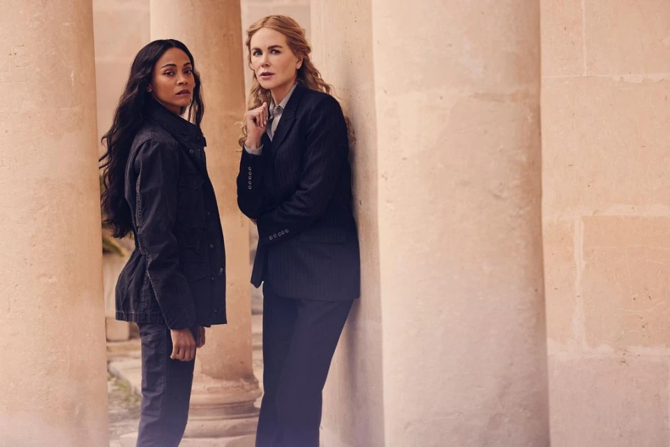 Two women, Zoe Saldana and Nicole Kidman, stand near stone columns, wearing dark jackets and pants, neutral expressions, in a medium outdoor shot.