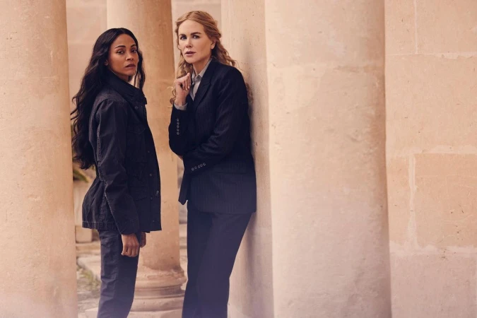 Two women, Zoe Saldana and Nicole Kidman, stand near stone columns, wearing dark jackets and pants, neutral expressions, in a medium outdoor shot.