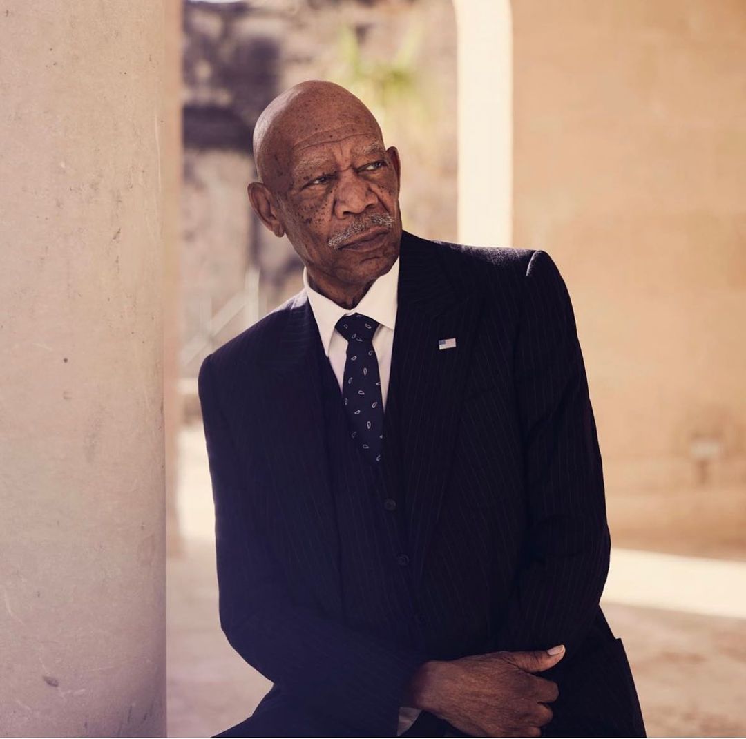 Portrait of a mature adult Black man in a dark suit and tie, standing near a concrete pillar, looking slightly to the side.
