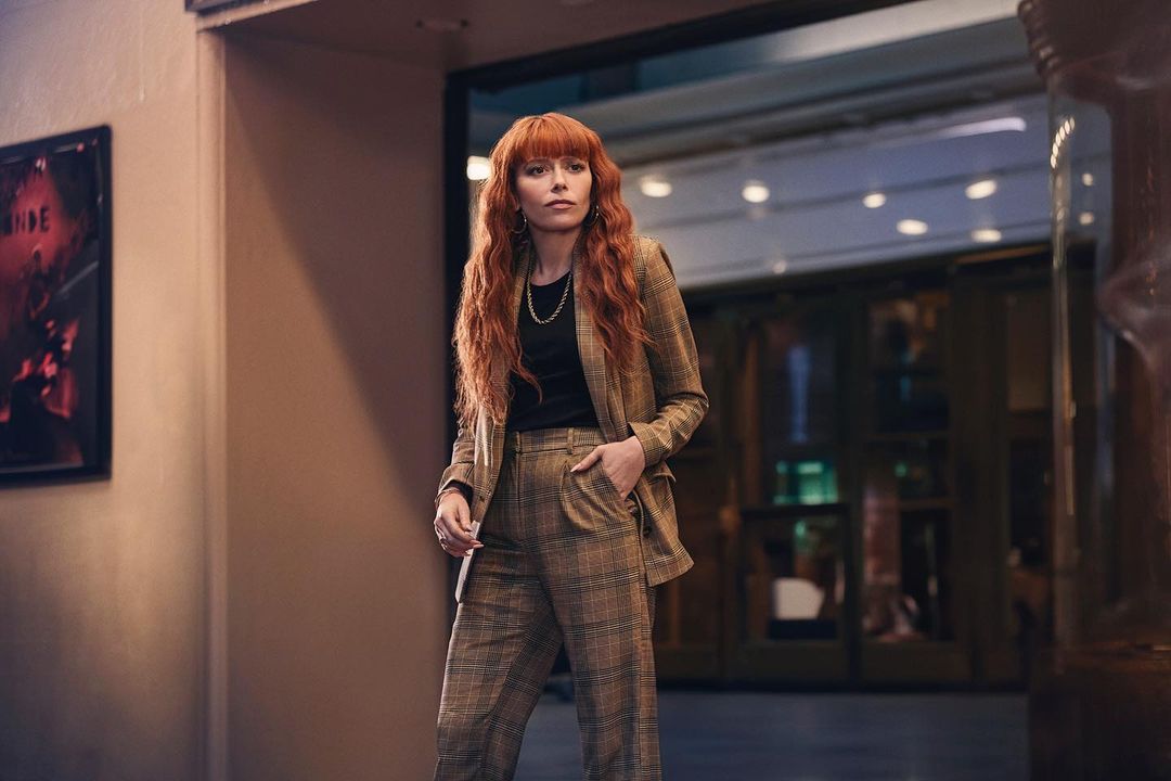 Stylish woman with red hair in plaid suit standing in hallway. Plaid suit and red hair are dominant features. Natural lighting.