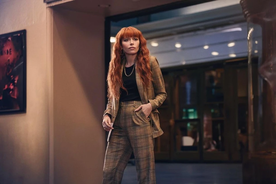 Stylish woman with red hair in plaid suit standing in hallway. Plaid suit and red hair are dominant features. Natural lighting.