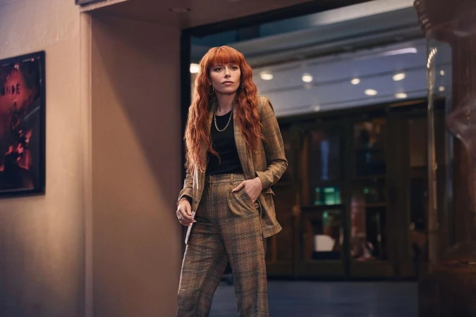 Stylish woman with red hair in plaid suit standing in hallway. Plaid suit and red hair are dominant features. Natural lighting.