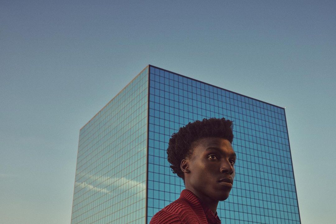 Portrait of a young Black man against a blue building and sky. He is wearing a textured red shirt. The building is square.