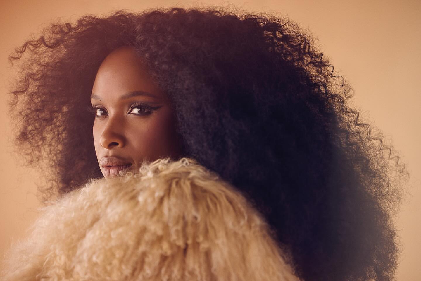 Portrait of a Black woman with dark curly hair, wearing a light-colored fur jacket, posing in front of a light tan studio backdrop.