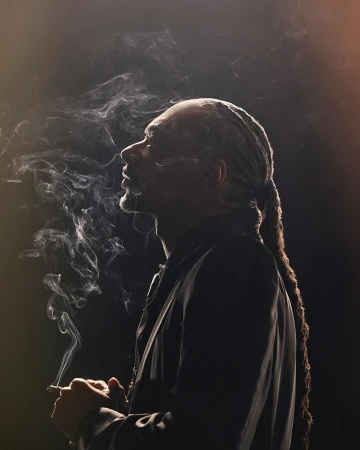 A side profile portrait of a man with dreadlocks, smoking, set against a dark background with soft lighting and atmospheric smoke.