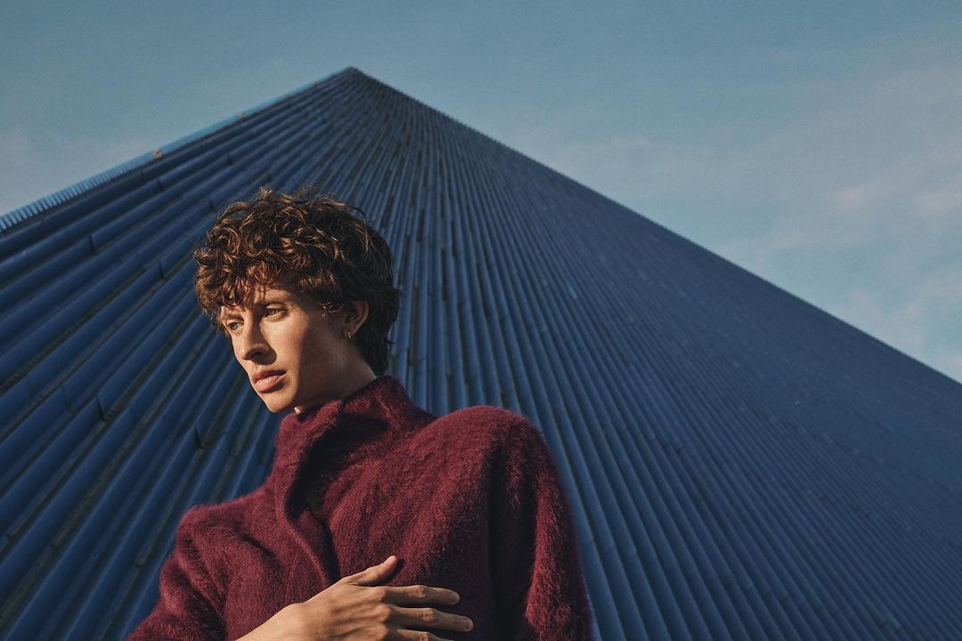 Man with curly hair in burgundy coat poses outdoors with blue corrugated building and sky backdrop for fashion or lifestyle content.
