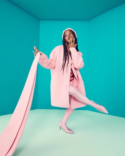 A woman in a pink coat and matching tights strikes a pose in a minimalist room with a turquoise background, wearing a headscarf.