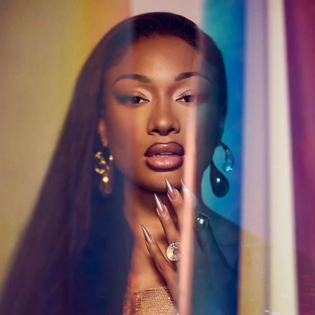 Portrait of a woman with long black hair wearing gold jewelry and a gold top posing in front of a multicolored background.