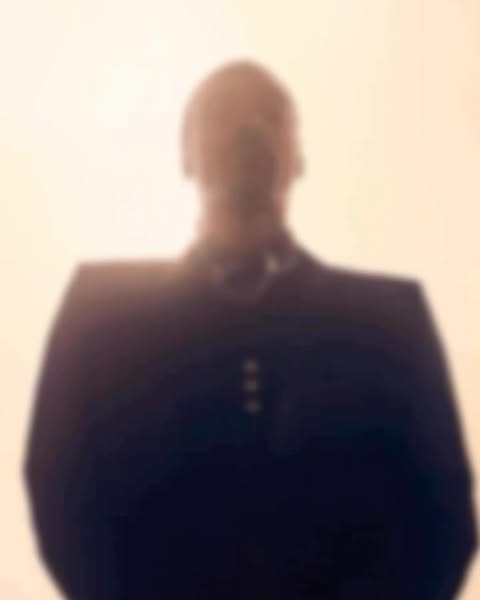Blurred portrait of an adult male wearing a suit jacket and white shirt, standing indoors against a light backdrop.