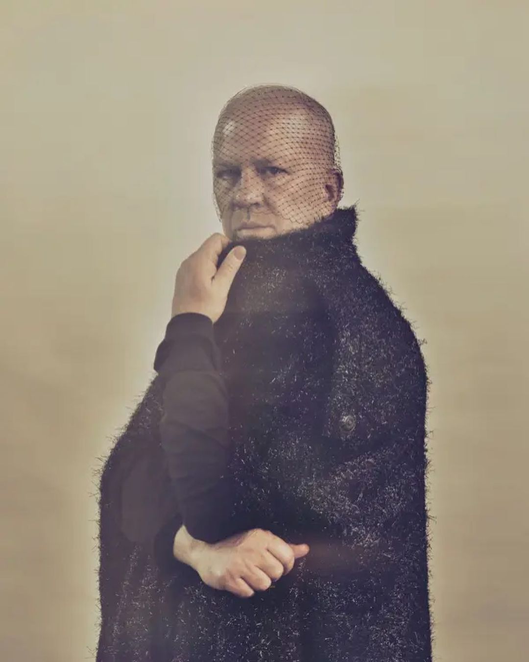 Portrait of a man wearing a mesh veil over his face and a textured black coat, photographed by Ramona Rosales.