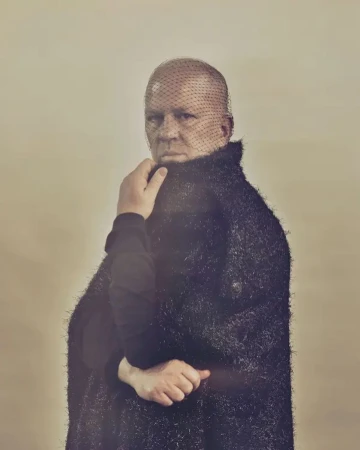 Portrait of a man wearing a mesh veil over his face and a textured black coat, photographed by Ramona Rosales.