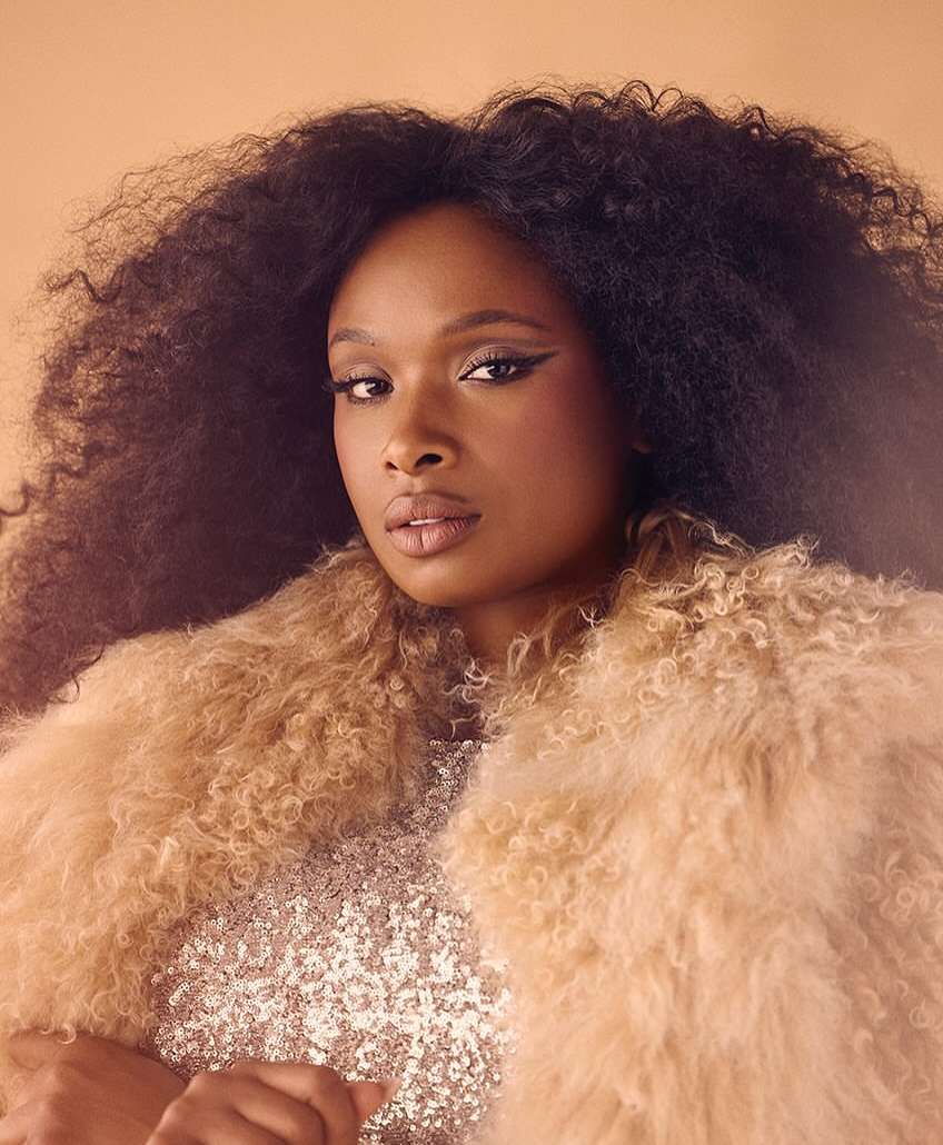 Close-up portrait of a Black woman with dark hair wearing a sequined top and a tan faux fur coat against a peach-colored backdrop.