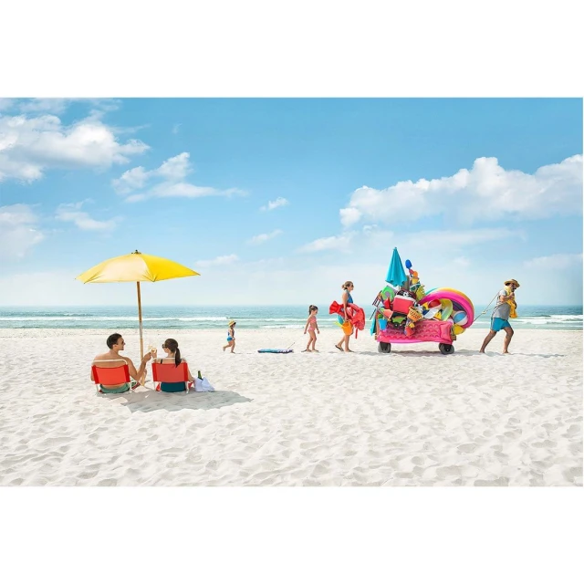 Family enjoying summer vacation at beach with a beach umbrella, chairs, and cart loaded with beach gear under blue sky.