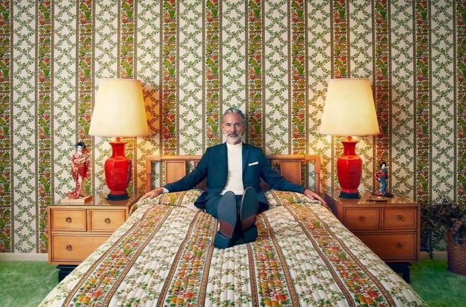 Middle-aged man in a suit sits on a bed with a floral quilt in a room with retro floral wallpaper, lamps, and nightstands.
