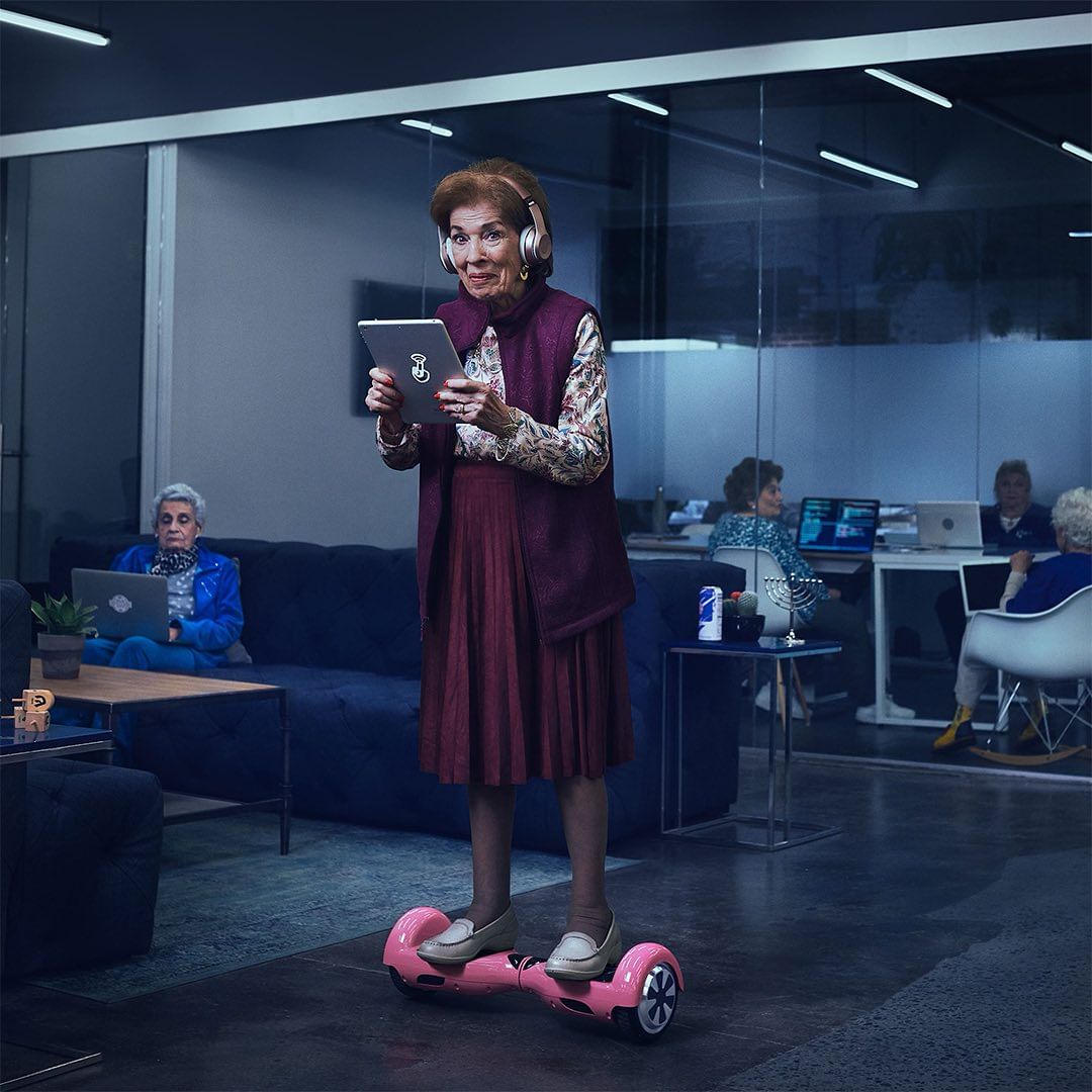 Elderly woman on hoverboard in modern office, wearing headphones, looking at tablet, with other seniors working in background.