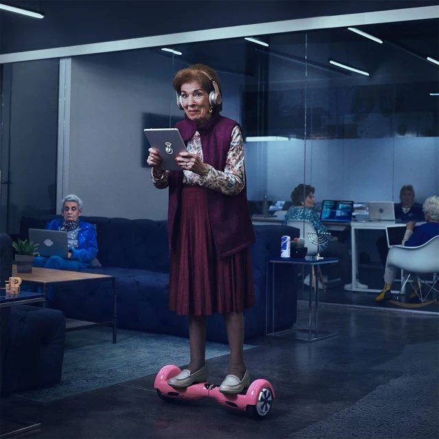 Elderly woman on hoverboard in modern office, wearing headphones, looking at tablet, with other seniors working in background.