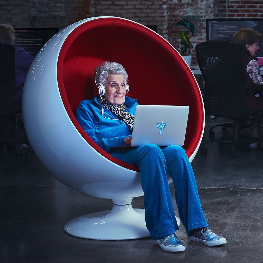 Elderly woman in a modern spherical chair wearing headphones using a laptop in a modern office space with other people.