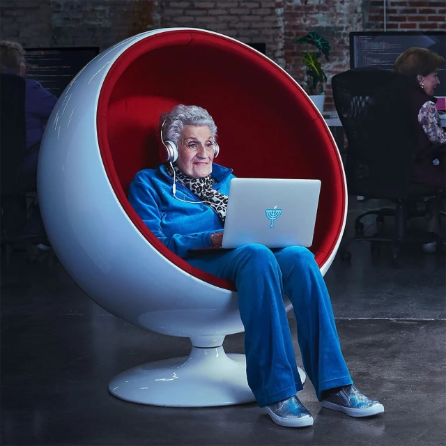 Elderly woman in a modern spherical chair wearing headphones using a laptop in a modern office space with other people.