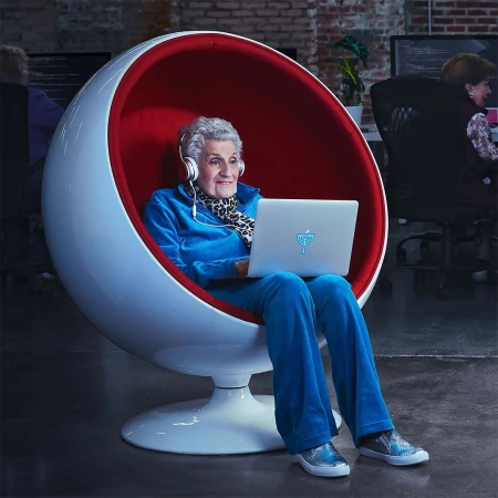 Elderly woman in a modern spherical chair wearing headphones using a laptop in a modern office space with other people.