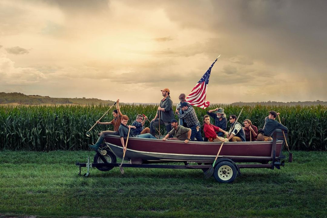 Group of people riding in a maroon boat on a trailer in a field, flying an American flag, near a cornfield under a cloudy sky.