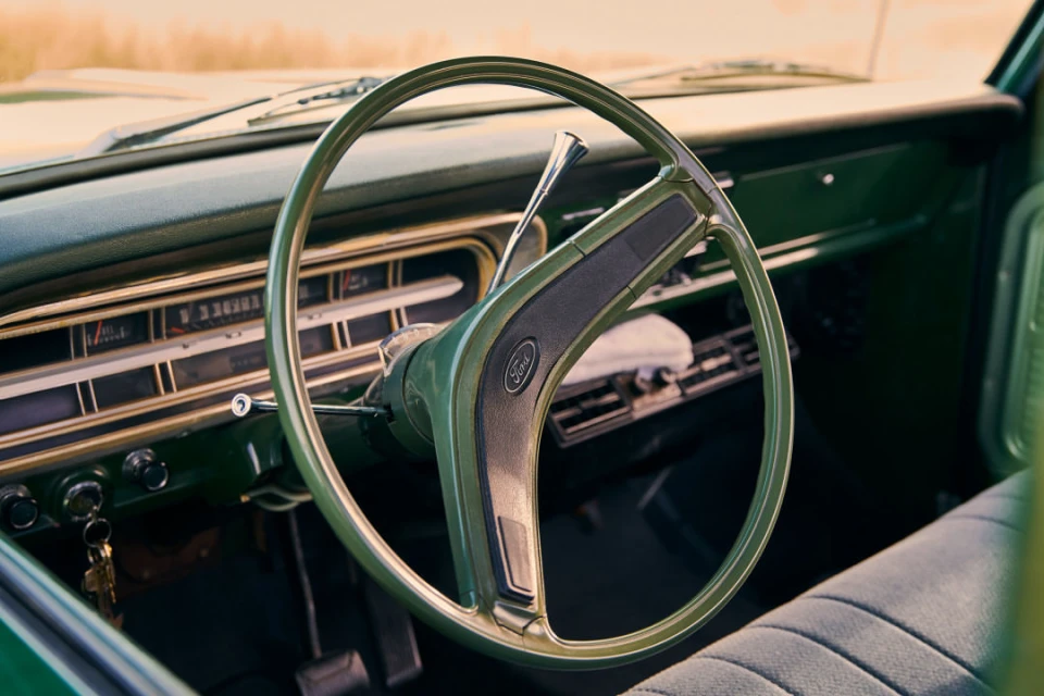Vintage Ford truck interior featuring a green steering wheel, dashboard, and a glimpse of the textured grey seat in daylight.