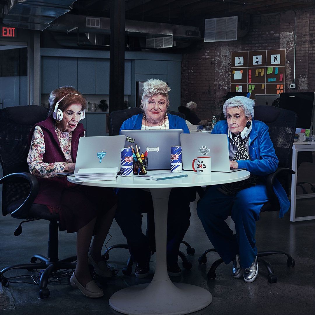 Three elderly women work at laptops around a white table in a modern office space. The scene is brightly lit with a brick wall background.