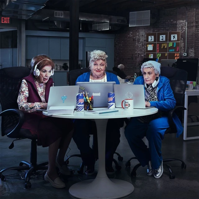 Three elderly women work at laptops around a white table in a modern office space. The scene is brightly lit with a brick wall background.