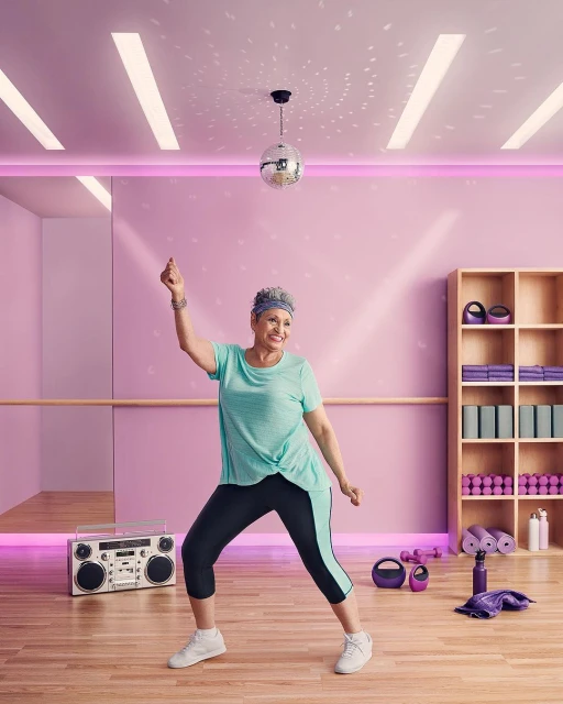 An older woman in a teal shirt and black leggings dances in a studio with a disco ball, boombox, and workout equipment.