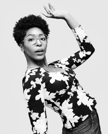 Black and white portrait of a black woman wearing patterned top and glasses with arm raised