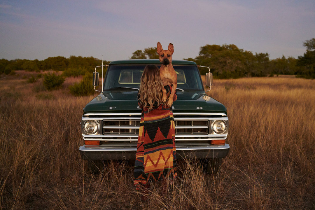 Woman and dog embrace by vintage Ford pickup truck in a field of tall grass, warm lighting, outdoor setting, casual fashion.