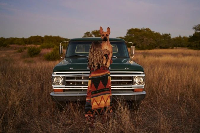 Woman and dog embrace by vintage Ford pickup truck in a field of tall grass, warm lighting, outdoor setting, casual fashion.