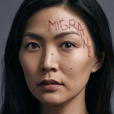 Portrait of an Asian woman with dark hair, a close-up shot with 'MIGRAINE' inscribed on her forehead, against a neutral gray background.
