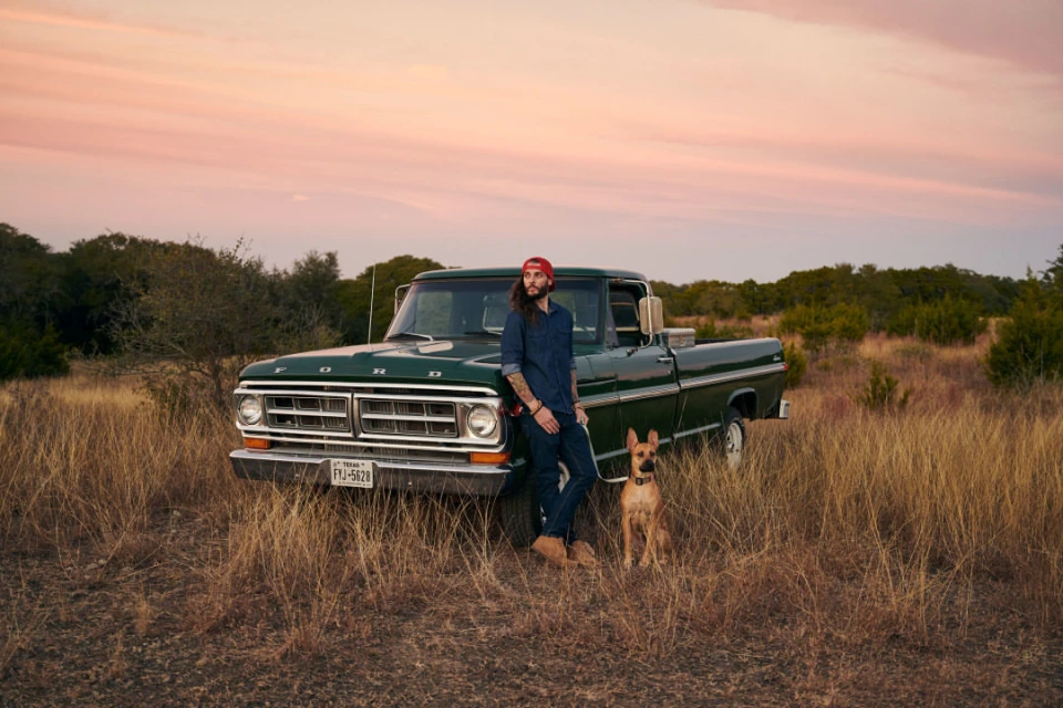 A man leans against a vintage green Ford pickup truck with his dog in a golden field under a pastel sunset sky.