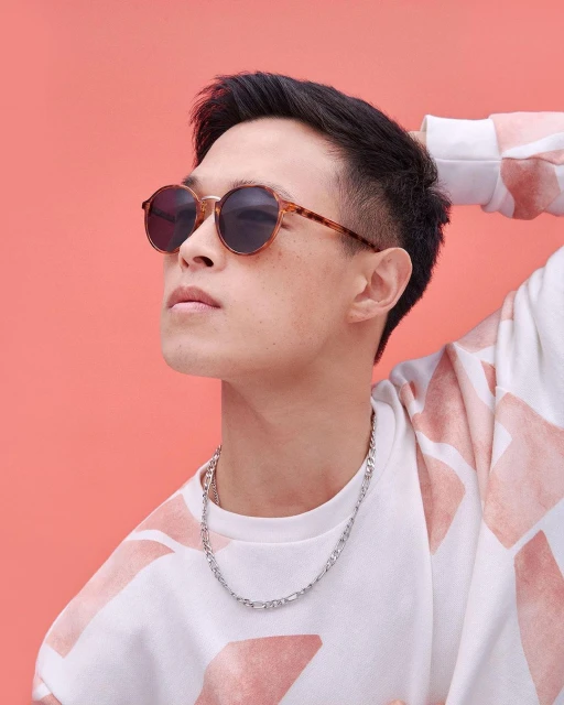 Portrait of a young adult male with a geometric patterned shirt, silver chain necklace, and round sunglasses against a peach background.