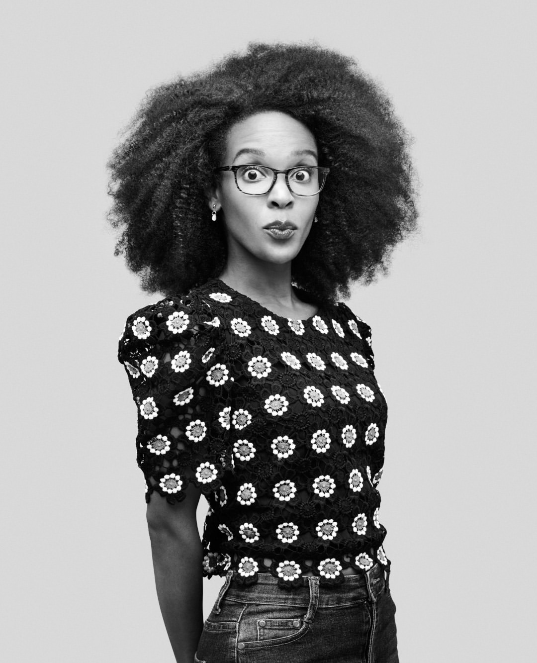 A young Black woman with an afro hairstyle and glasses makes a surprised face while wearing a floral blouse against a gray backdrop.