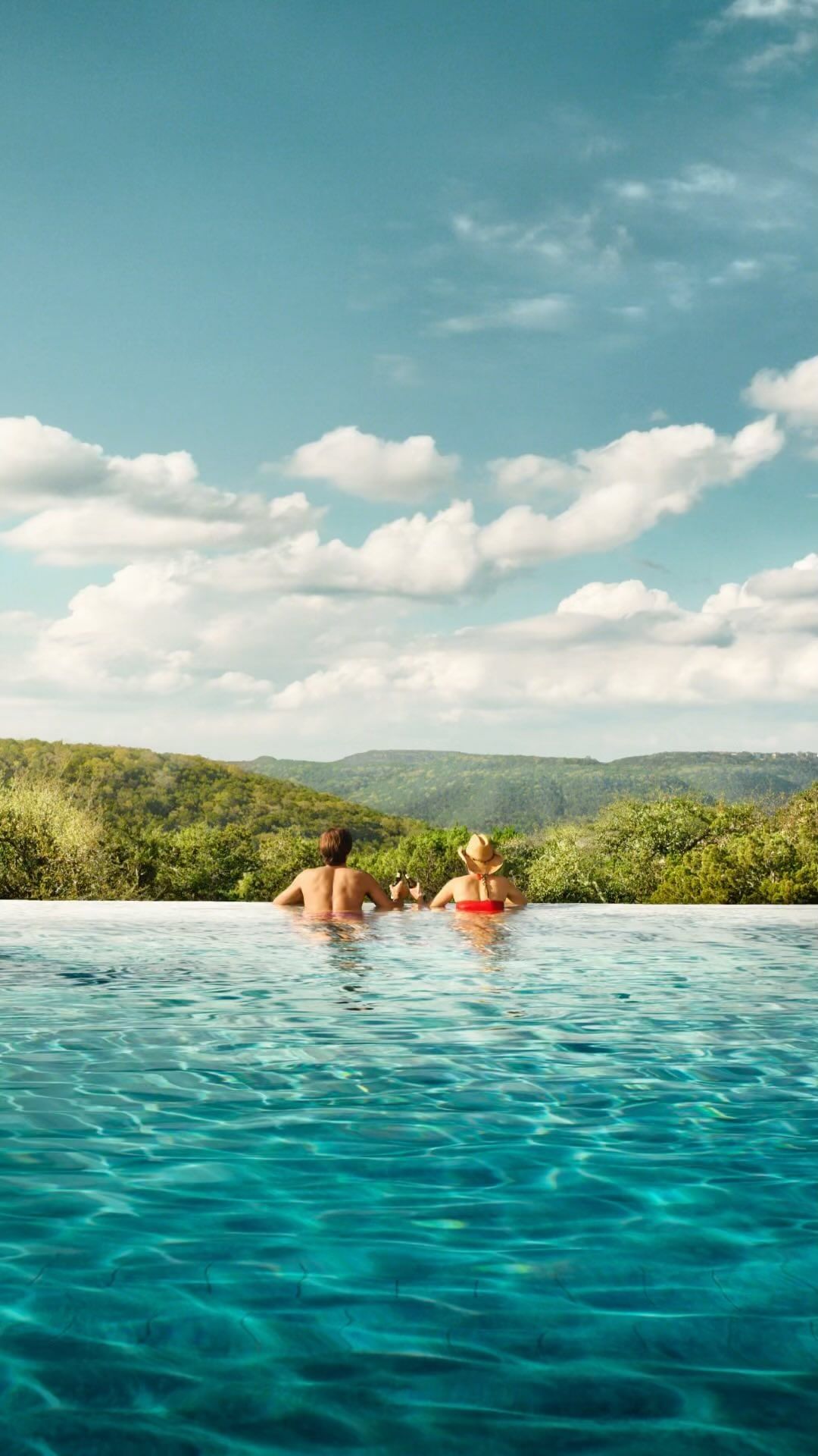 Two people enjoy the view from an infinity pool overlooking a lush green landscape under a partly cloudy sky, creating a serene travel scene.