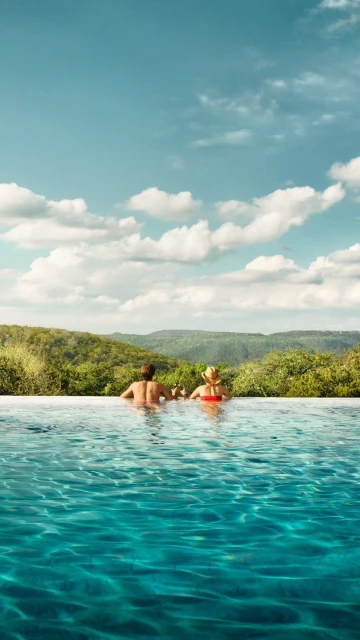 Two people enjoy the view from an infinity pool overlooking a lush green landscape under a partly cloudy sky, creating a serene travel scene.