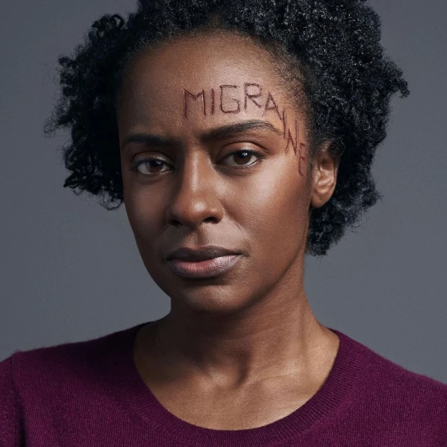 Close-up portrait of a dark-skinned adult female with curly hair, wearing a maroon sweater, with 'MIGRAINE' written on her forehead.