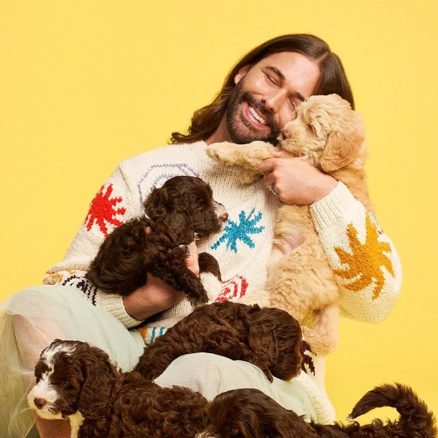 Jonathan Van Ness smiles while holding a golden doodle puppy, surrounded by brown and white puppies against a yellow background.