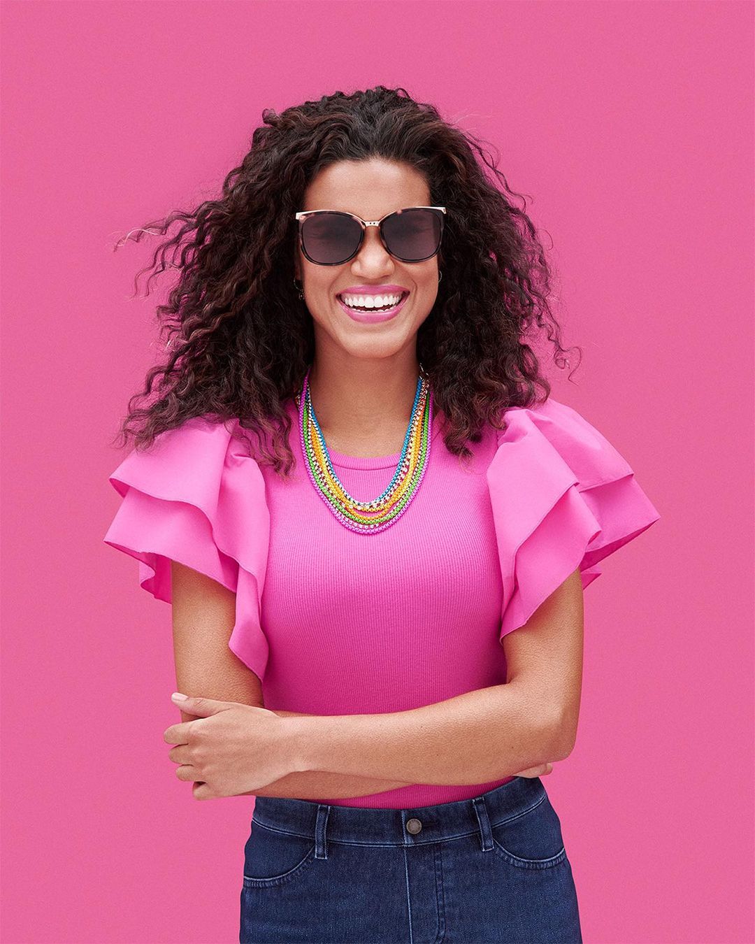 Smiling woman in pink top and sunglasses stands against pink backdrop, showcasing a vibrant layered necklace in a medium studio shot.
