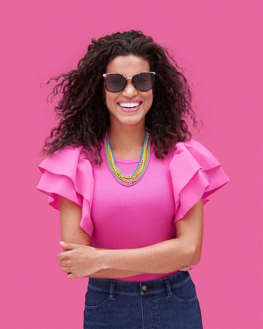 Smiling woman in pink top and sunglasses stands against pink backdrop, showcasing a vibrant layered necklace in a medium studio shot.
