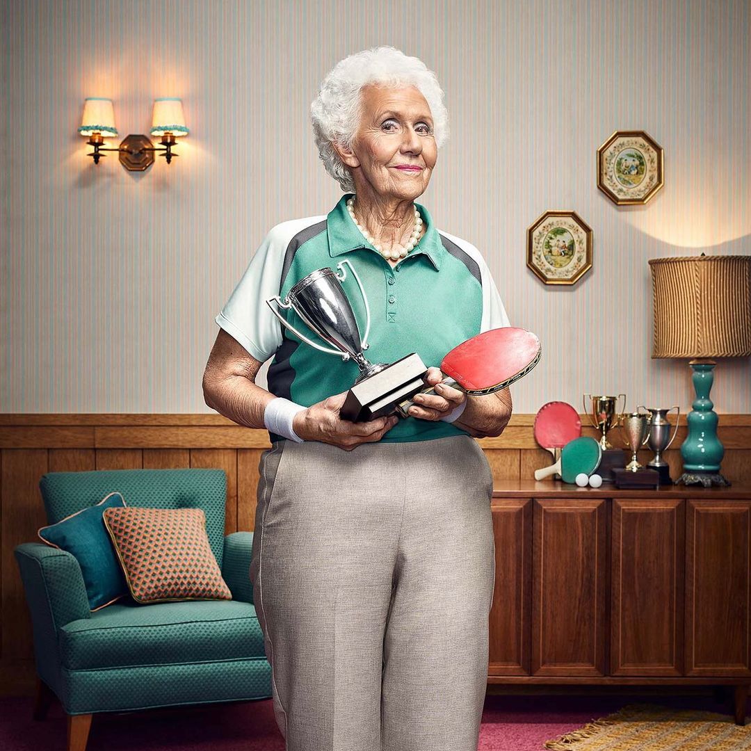 Elderly woman in sportswear holds a trophy and ping pong paddle in a retro-style indoor setting with vintage furniture.
