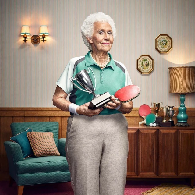 Elderly woman in sportswear holds a trophy and ping pong paddle in a retro-style indoor setting with vintage furniture.