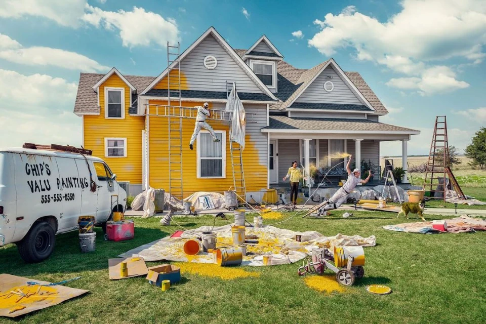 House being painted by a group of people using ladders and sprayers, with paint cans, paintbrushes, and tarps scattered around the lawn.