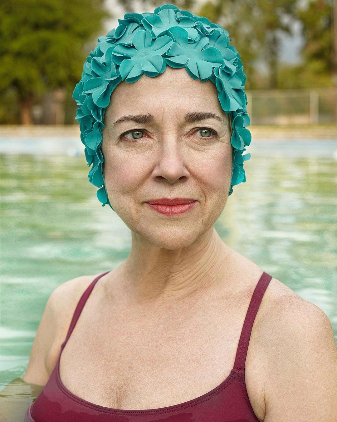 Adult woman with a turquoise flower swim cap at the edge of a pool with a blurry outdoor background, shot with a shallow depth of field.