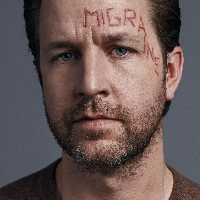 Close-up portrait of an adult male with light skin, blue eyes, and a beard, with the word 'MIGRAINE' graphically displayed on his face.