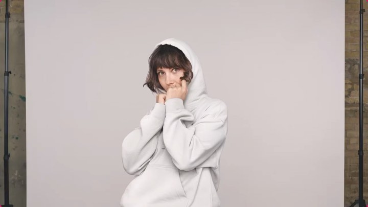 Portrait of a young adult woman in a hooded sweatshirt, covering her mouth, set against a neutral backdrop in a studio setting.