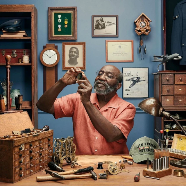 An elderly Black man wearing glasses and an orange shirt carefully examines a watch component in a workshop filled with tools and memorabilia.